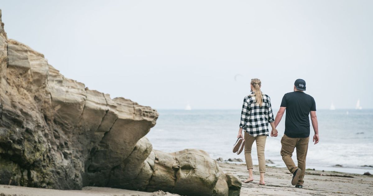 A couple holds hands walking along a California beach past coastal rocks, sailboats on the ocean behind them.