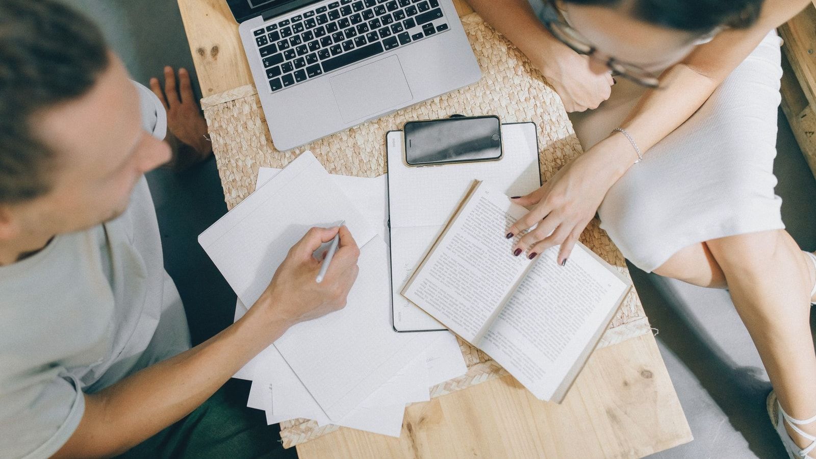 Overhead view of coworkers discussing ideas with laptop, papers, and books on a wooden desk.