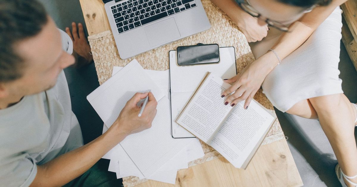 Overhead view of coworkers discussing ideas with laptop, papers, and books on a wooden desk.