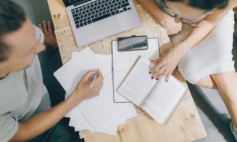 Overhead view of coworkers discussing ideas with laptop, papers, and books on a wooden desk.