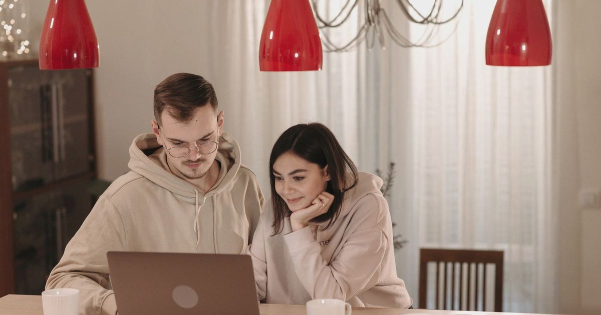 A couple in casual hoodies using a laptop at home, smiling and relaxed.