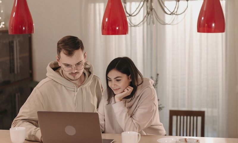 A couple in casual hoodies using a laptop at home, smiling and relaxed.