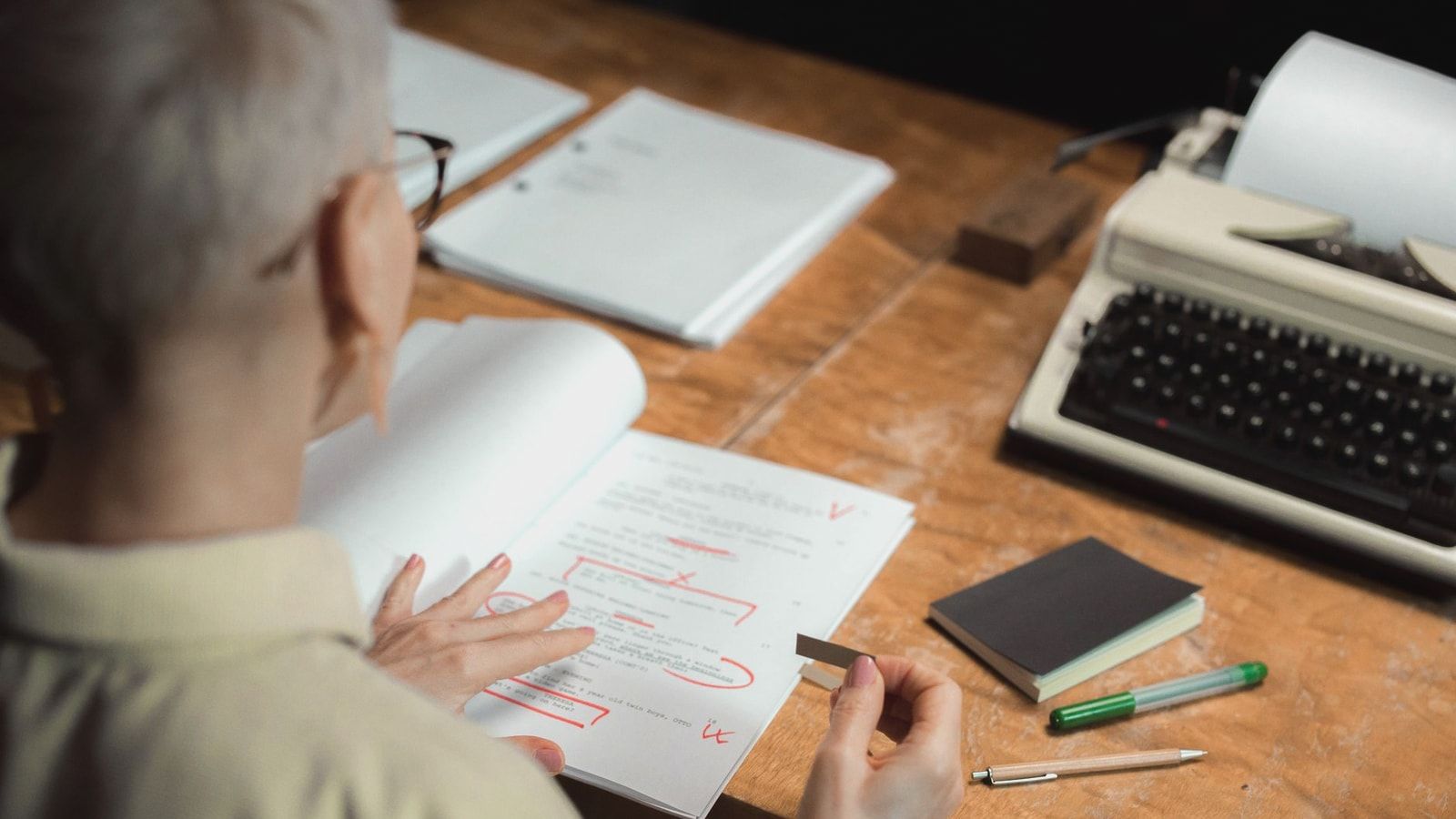 An adult woman reviewing a script with red pen marks at a wooden desk with a typewriter.