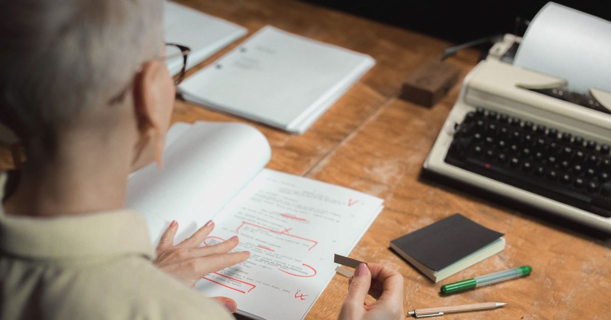 An adult woman reviewing a script with red pen marks at a wooden desk with a typewriter.