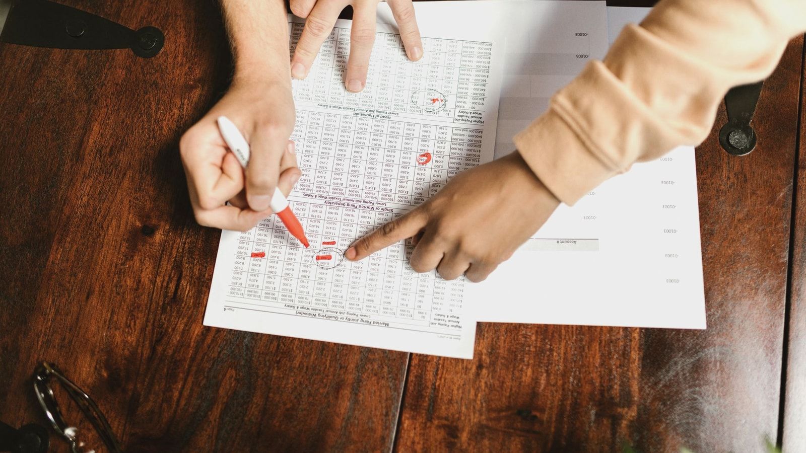 Two hands reviewing and marking financial documents with a red pen on a wooden table, top view.