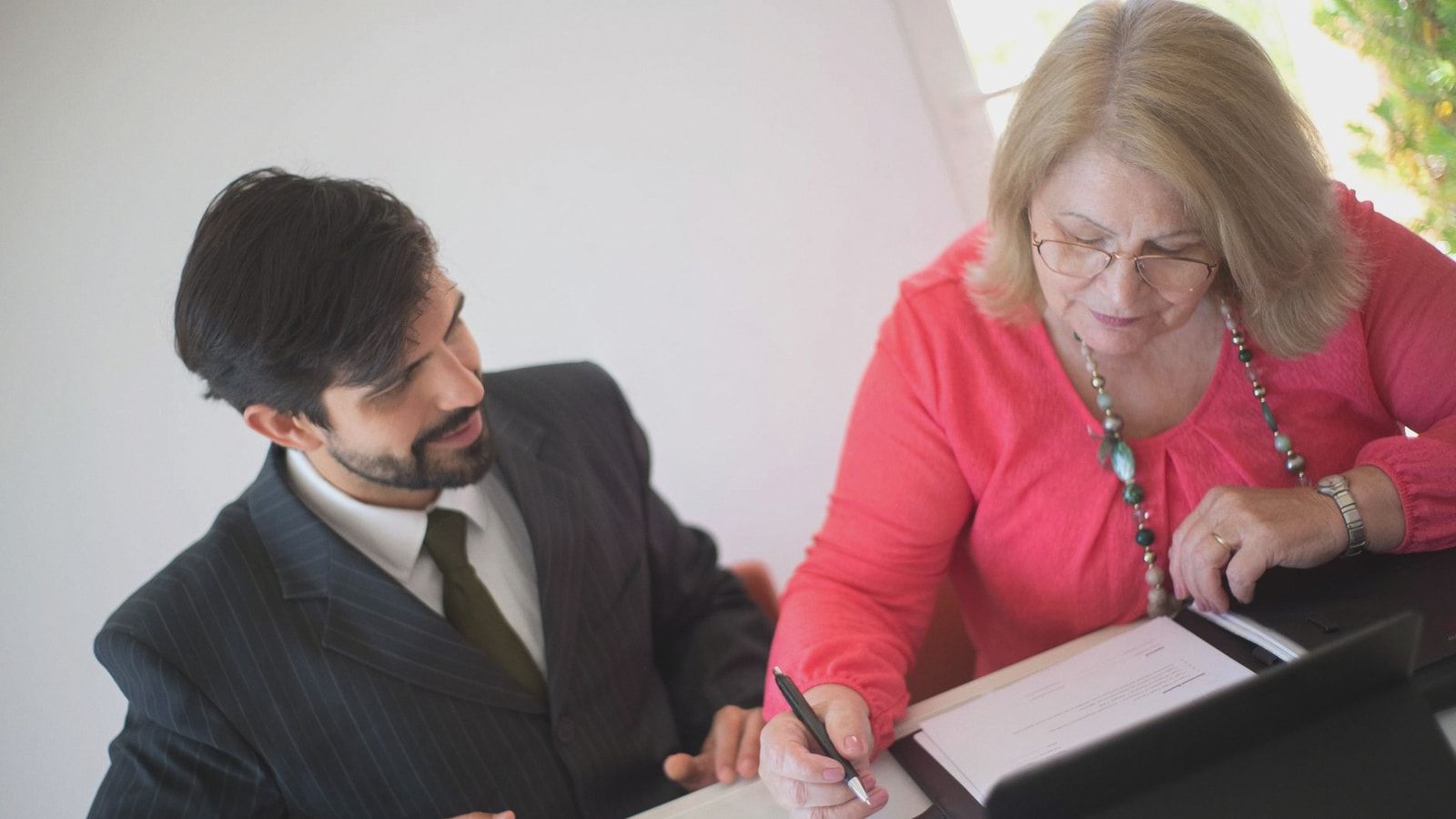 A senior woman signing paperwork with a young professional assisting her at a desk.