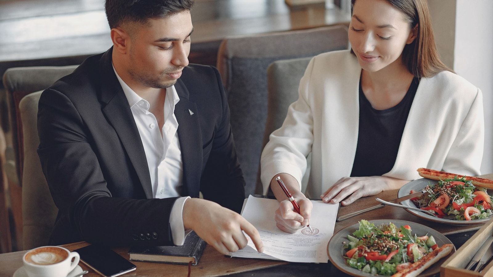 Concentrated multiethnic colleagues in formal clothes sitting at table in cafe with cup of espresso and plates of salad while signing documents