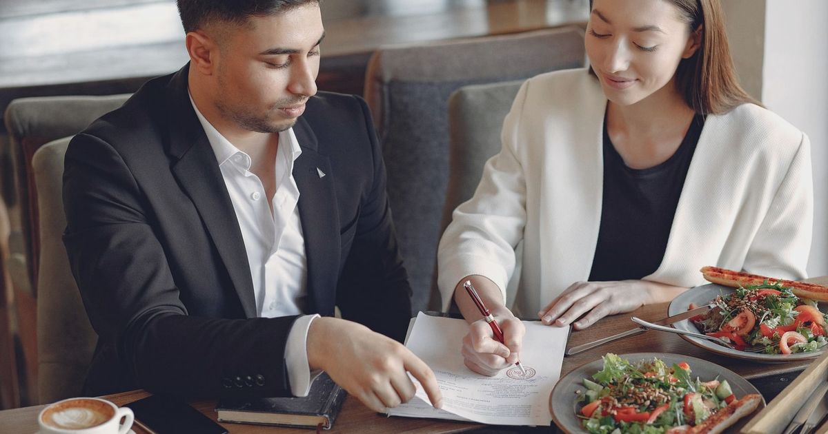 Concentrated multiethnic colleagues in formal clothes sitting at table in cafe with cup of espresso and plates of salad while signing documents