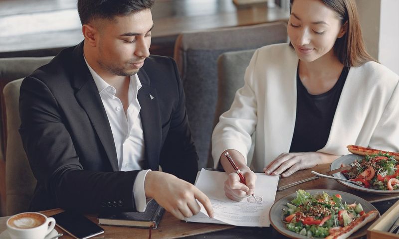 Concentrated multiethnic colleagues in formal clothes sitting at table in cafe with cup of espresso and plates of salad while signing documents
