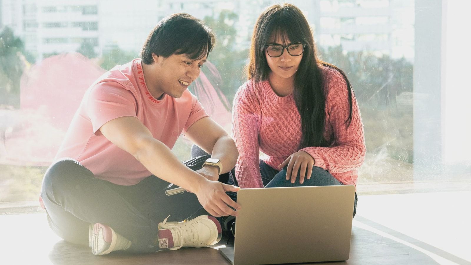 Two young adults in casual attire working together on a laptop by a window.