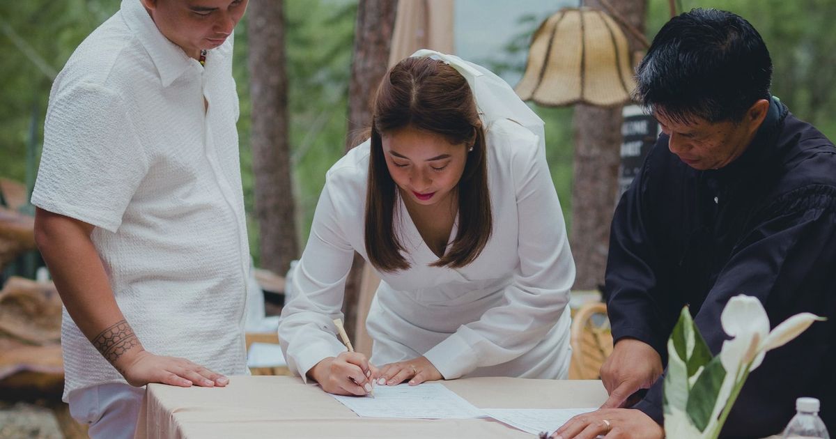 Bride signing marriage certificate surrounded by groom and officiant during outdoor wedding.