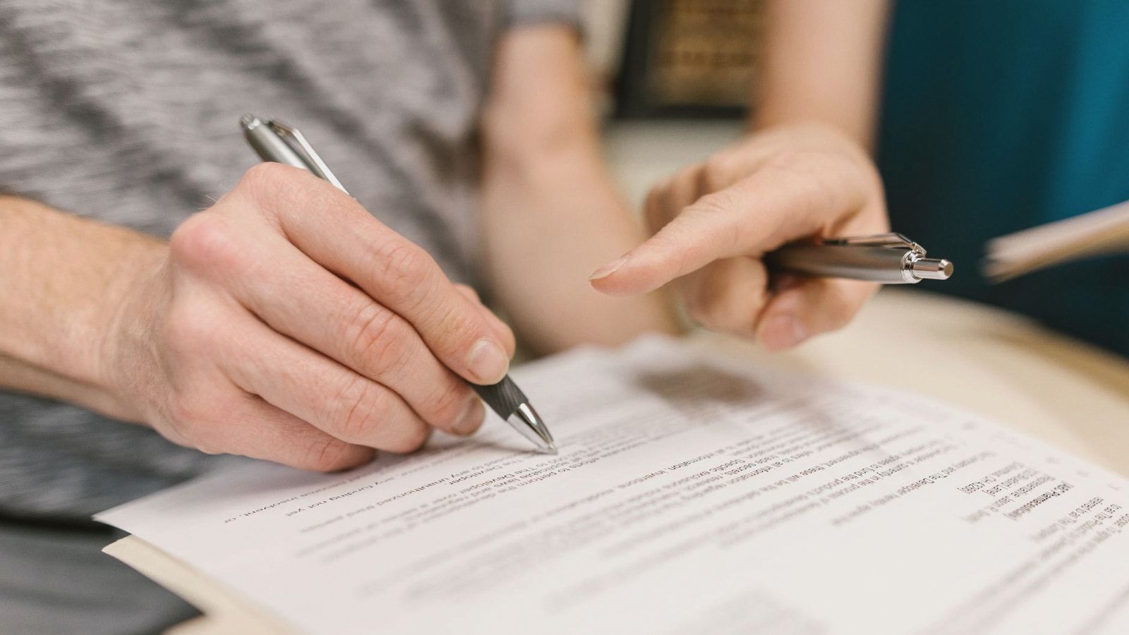 Close-up of two adults reviewing and signing a legal document with pens indoors.