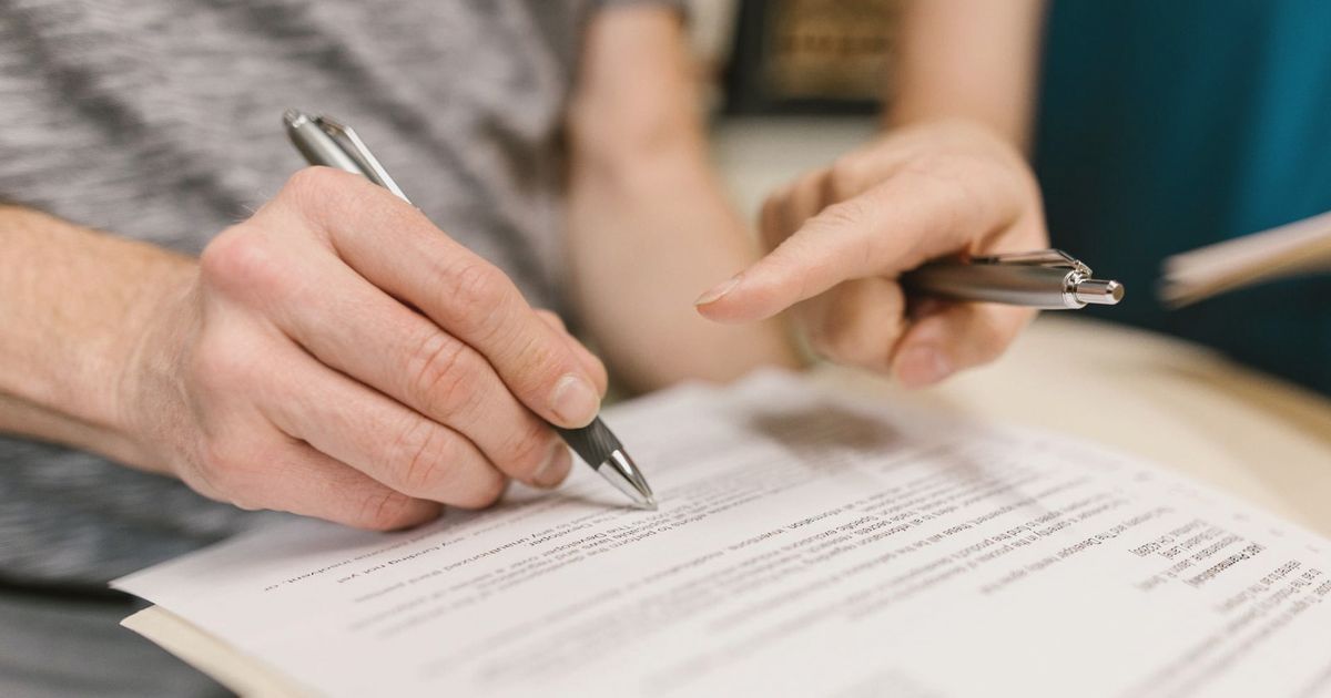 Close-up of two adults reviewing and signing a legal document with pens indoors.