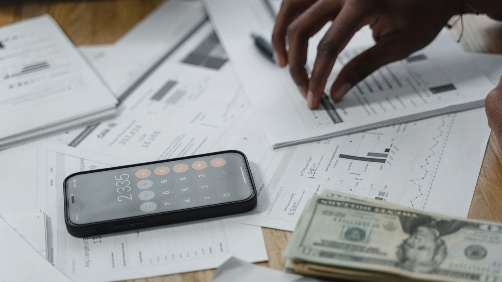 Close-up of hands calculating financial data with reports, smartphone, and cash on table.