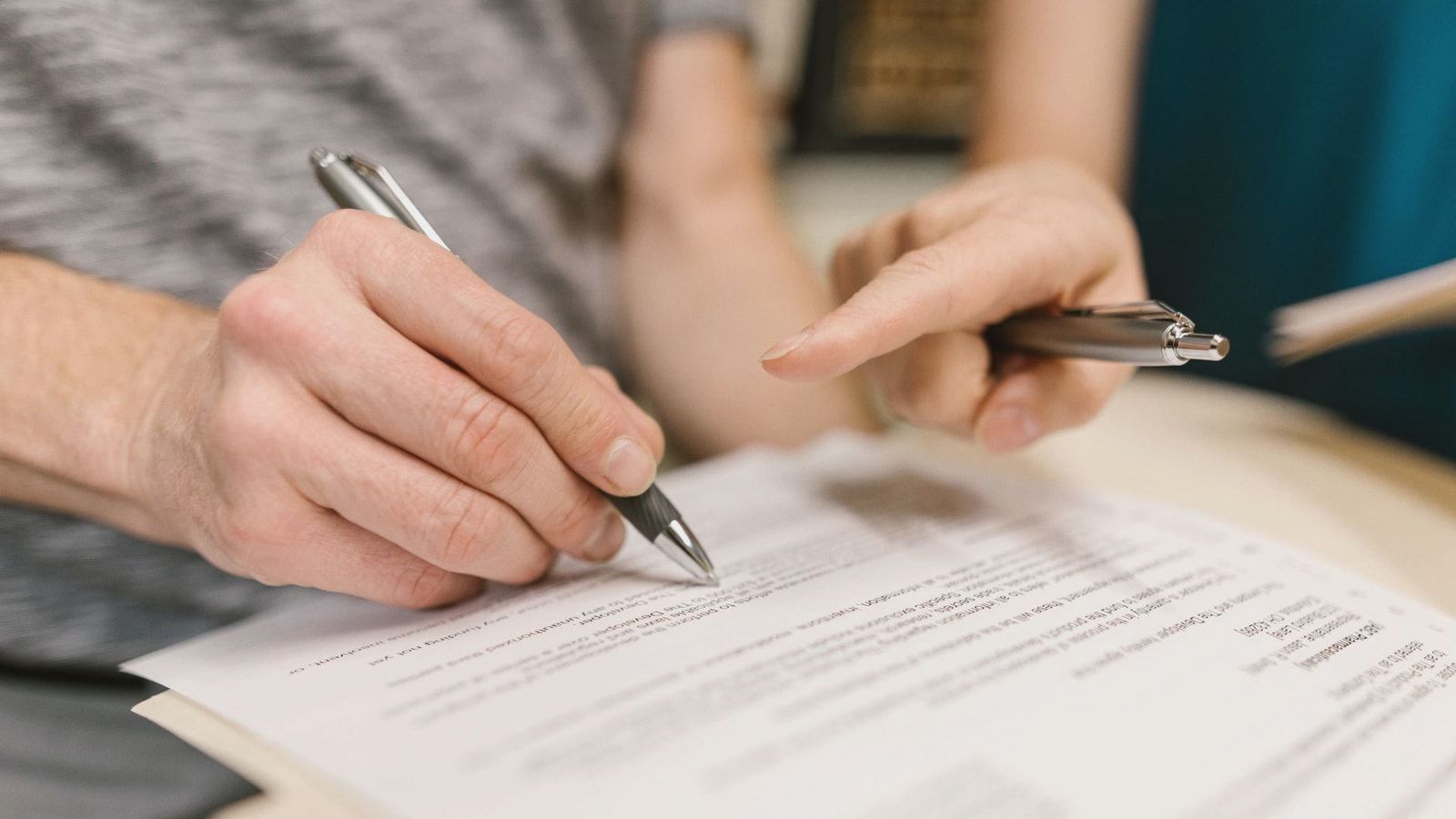 Close-up of two adults reviewing and signing a legal document with pens indoors.