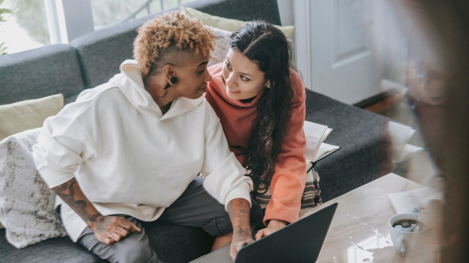 High angle of multiracial couple in casual clothes sitting close on couch with netbook and looking at each other