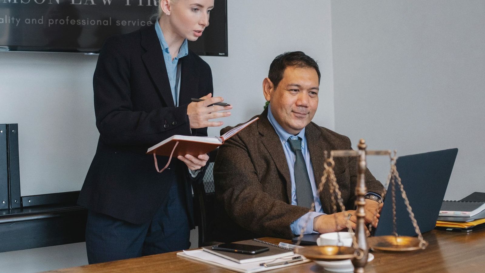 Concentrated young female assistant taking notes in planner while standing at table near middle aged ethnic male colleague typing on laptop during work in law firm