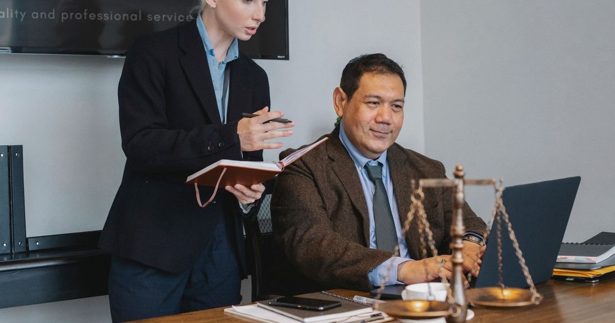 Concentrated young female assistant taking notes in planner while standing at table near middle aged ethnic male colleague typing on laptop during work in law firm