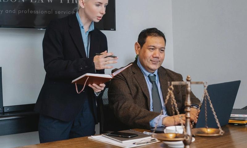 Concentrated young female assistant taking notes in planner while standing at table near middle aged ethnic male colleague typing on laptop during work in law firm
