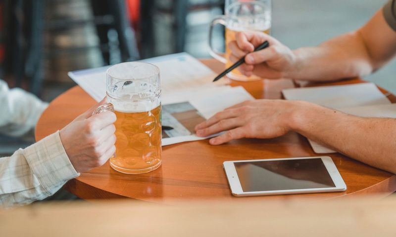 Side view of crop colleagues checking important papers near tablet and glasses of beer on table