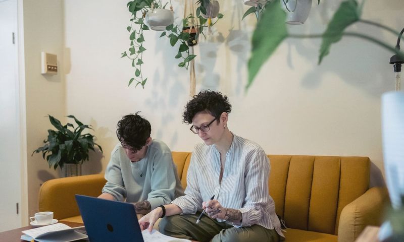 Concentrated young tattooed female freelancers with short dark hair in casual clothes sitting on comfortable sofa and working remotely on laptop
