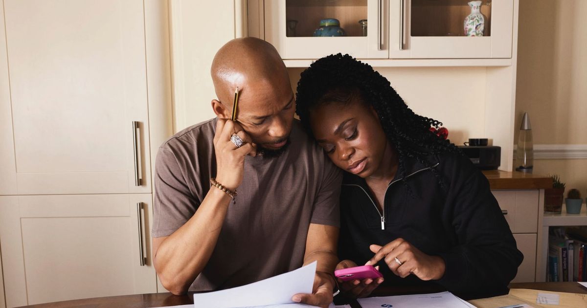 A couple sits at a table managing domestic finances, evaluating documents and using a smartphone.