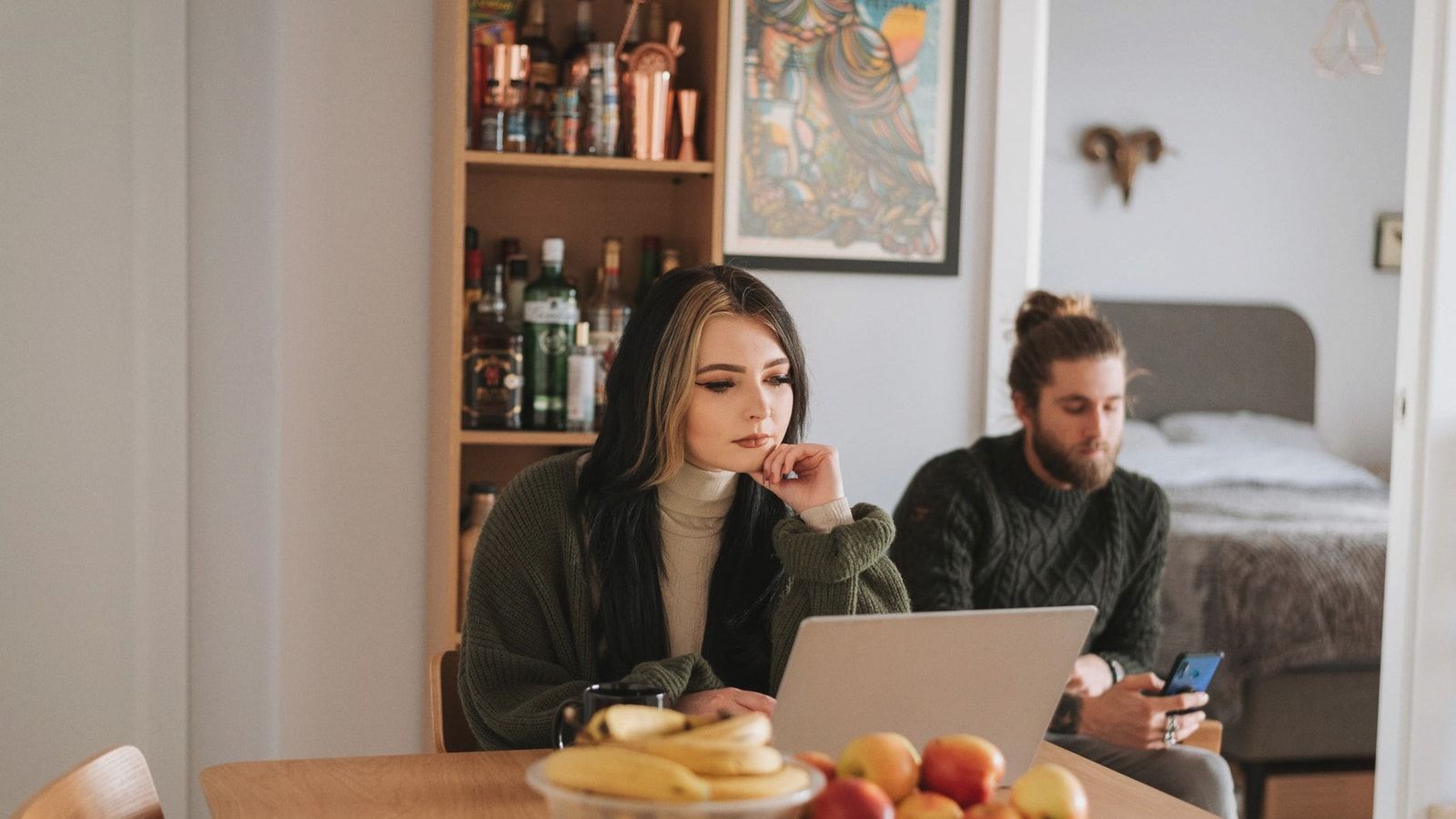Young woman surfing internet on netbook at table with assorted fruits against boyfriend chatting on cellphone at home