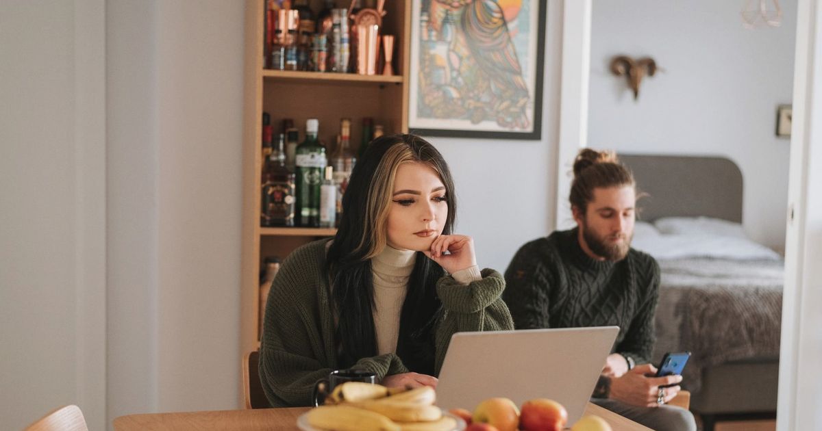 Young woman surfing internet on netbook at table with assorted fruits against boyfriend chatting on cellphone at home