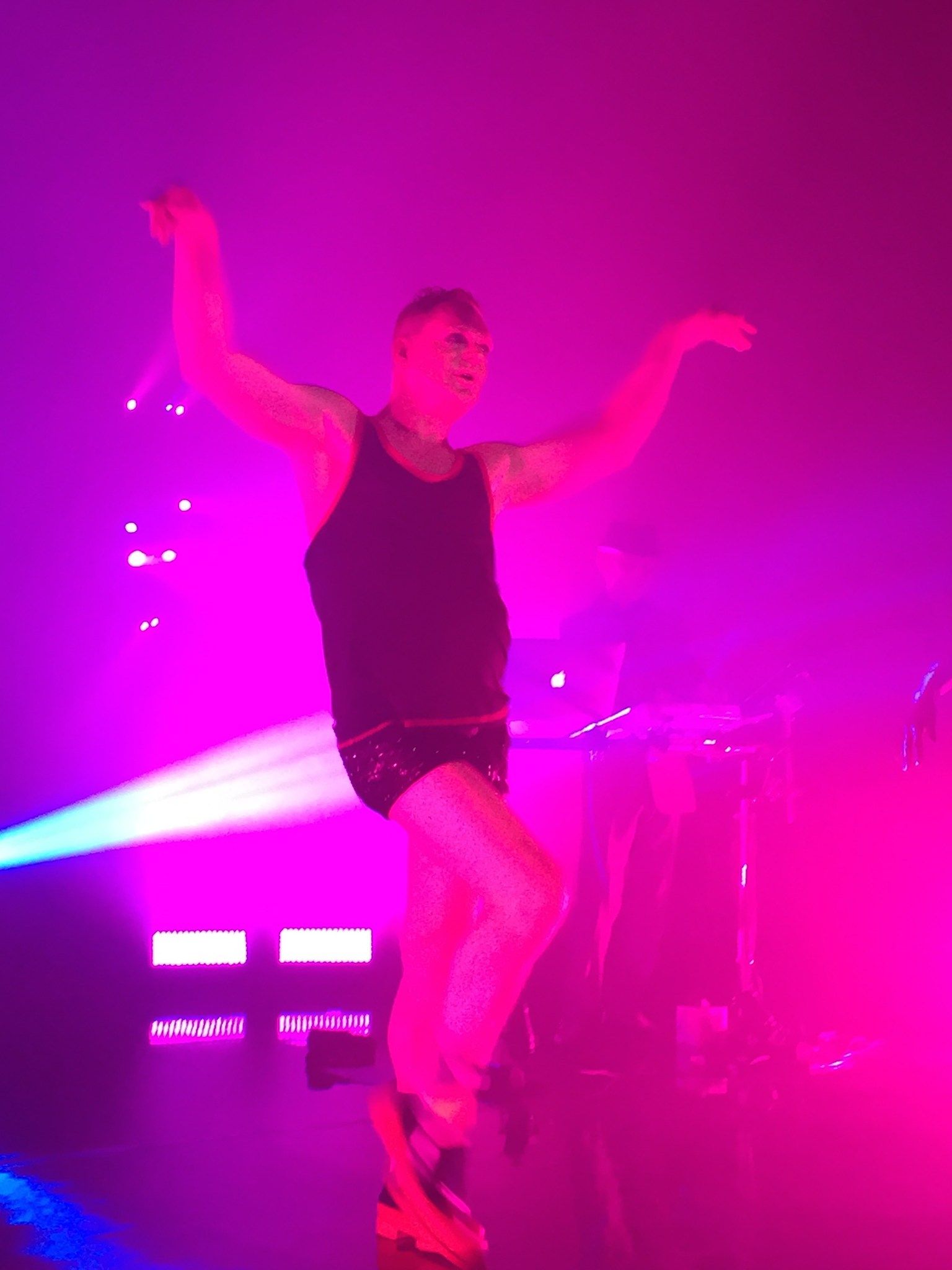 Andy Bell of Erasure dancing on stage in a vest and shorts, one knee raised, under pink stage lights