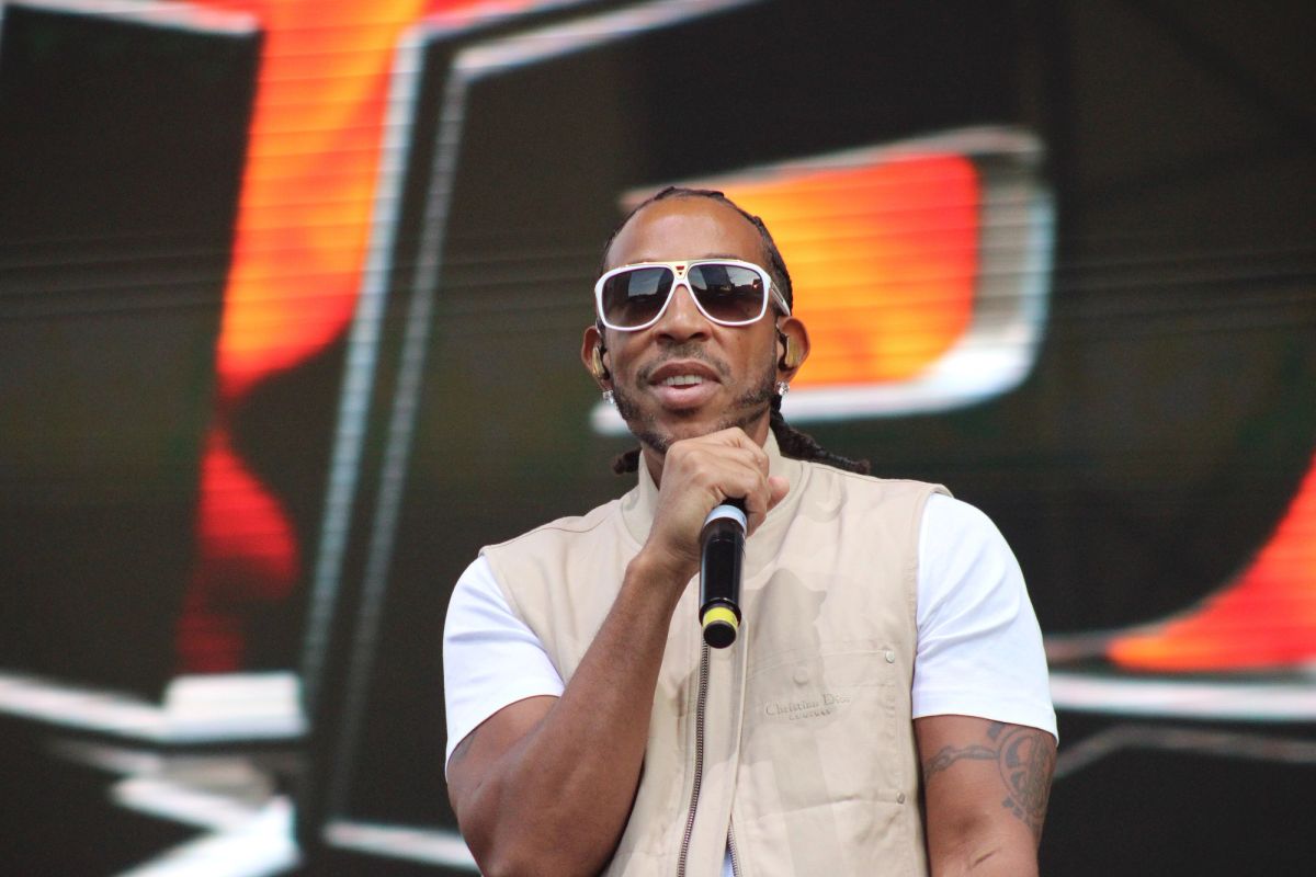 Ludacris holding a microphone on stage, wearing white sunglasses and a beige vest