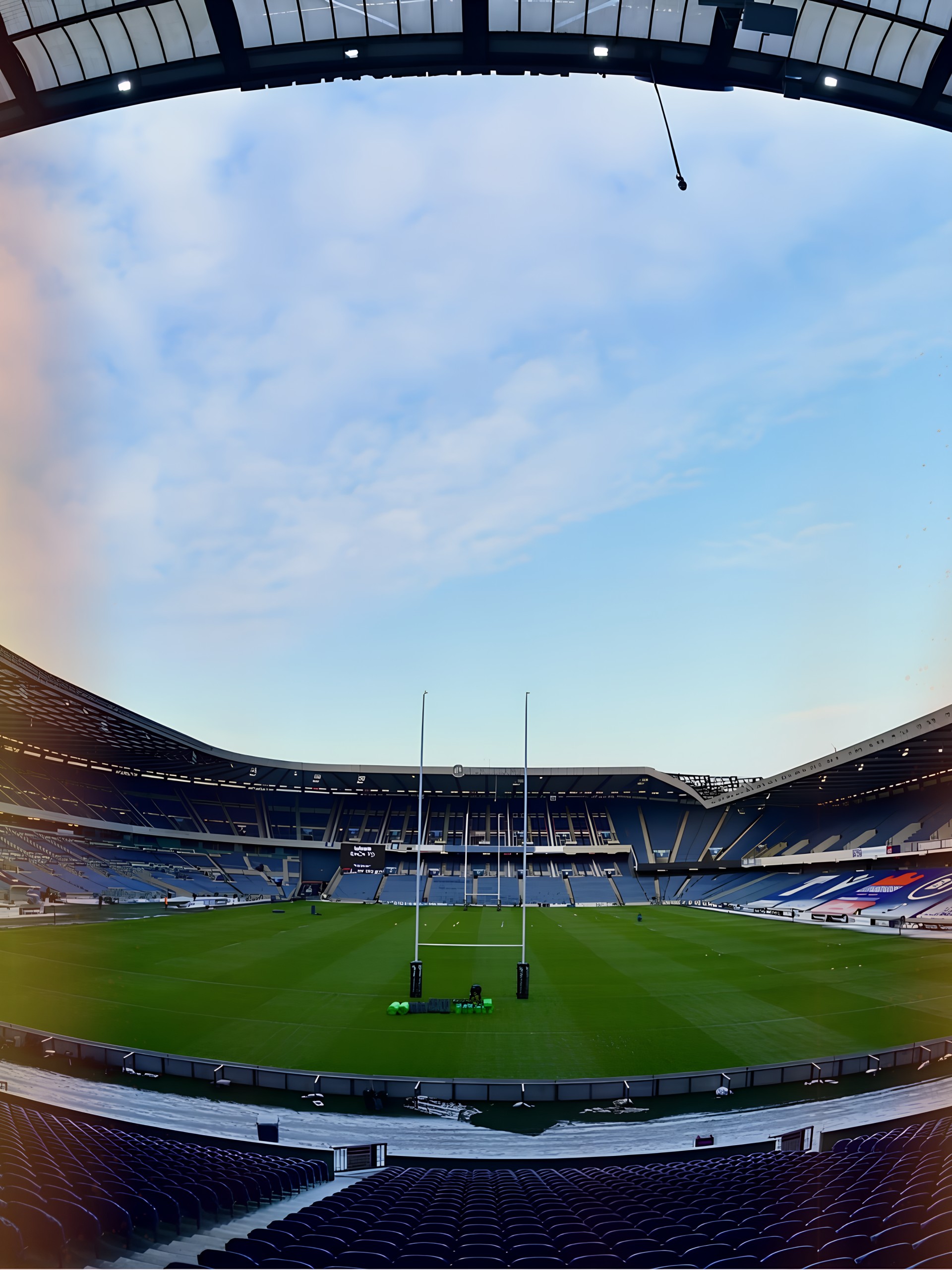 Empty rugby stadium seen from the stands, with blue seats, the posts and a green pitch under an evening sky