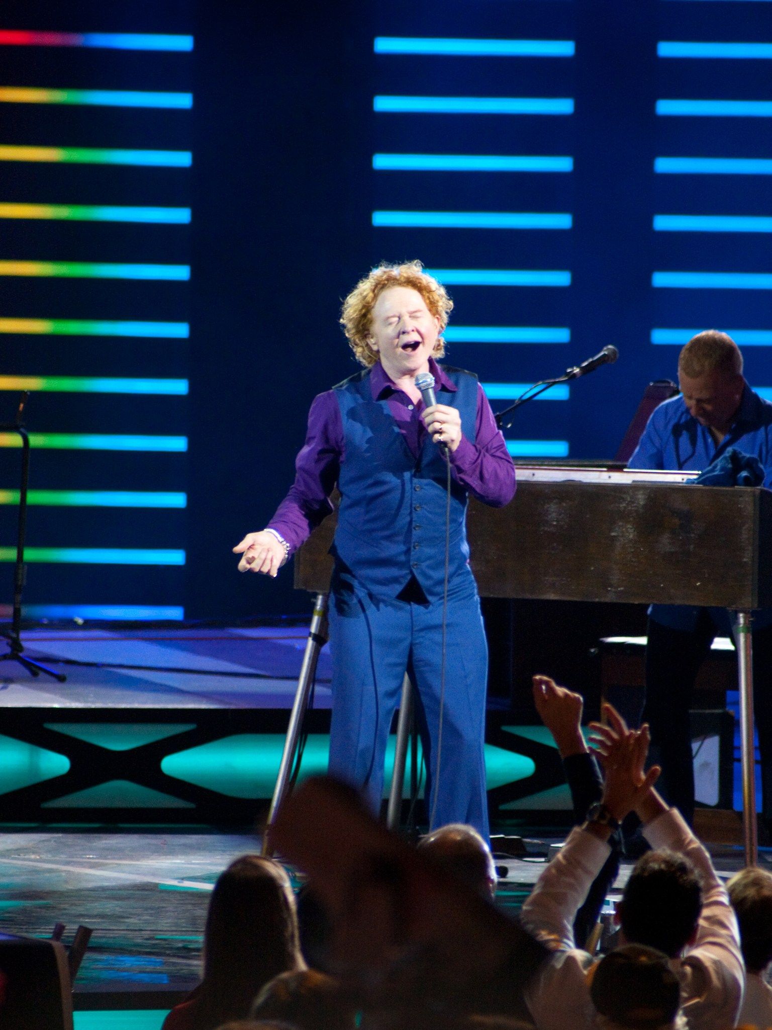 Mick Hucknall singing with his eyes closed on stage while fans clap in the foreground