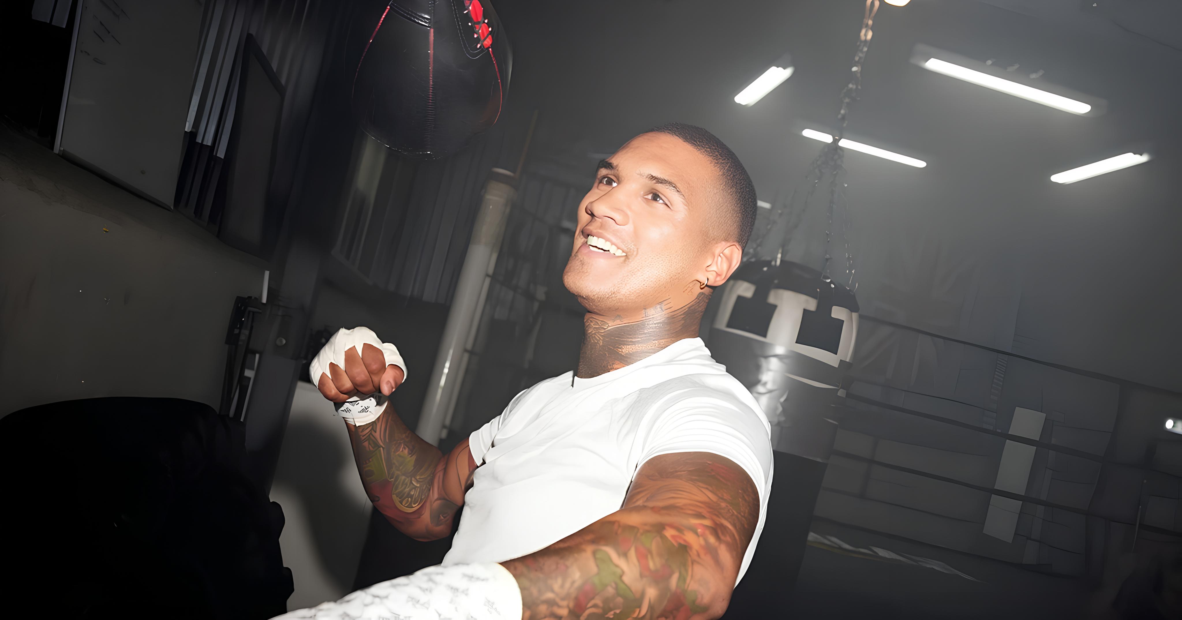 Conor Benn training in boxing gym, smiling, with tattoos, Union Jack flag, fluorescent lights.