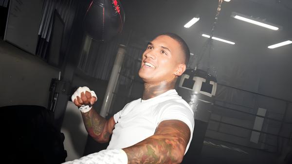 Conor Benn training in boxing gym, smiling, with tattoos, Union Jack flag, fluorescent lights.