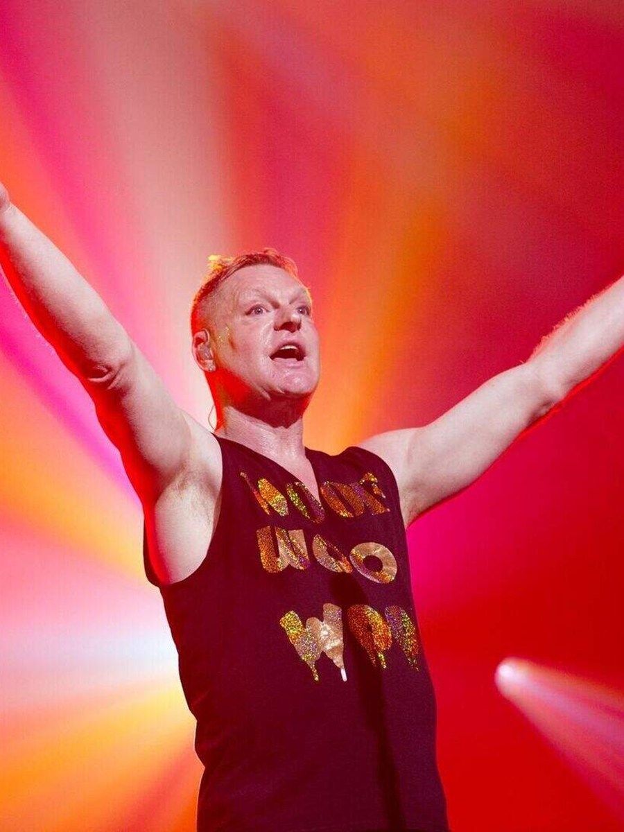 Andy Bell of Erasure singing on stage with his arms outstretched under red and orange lights