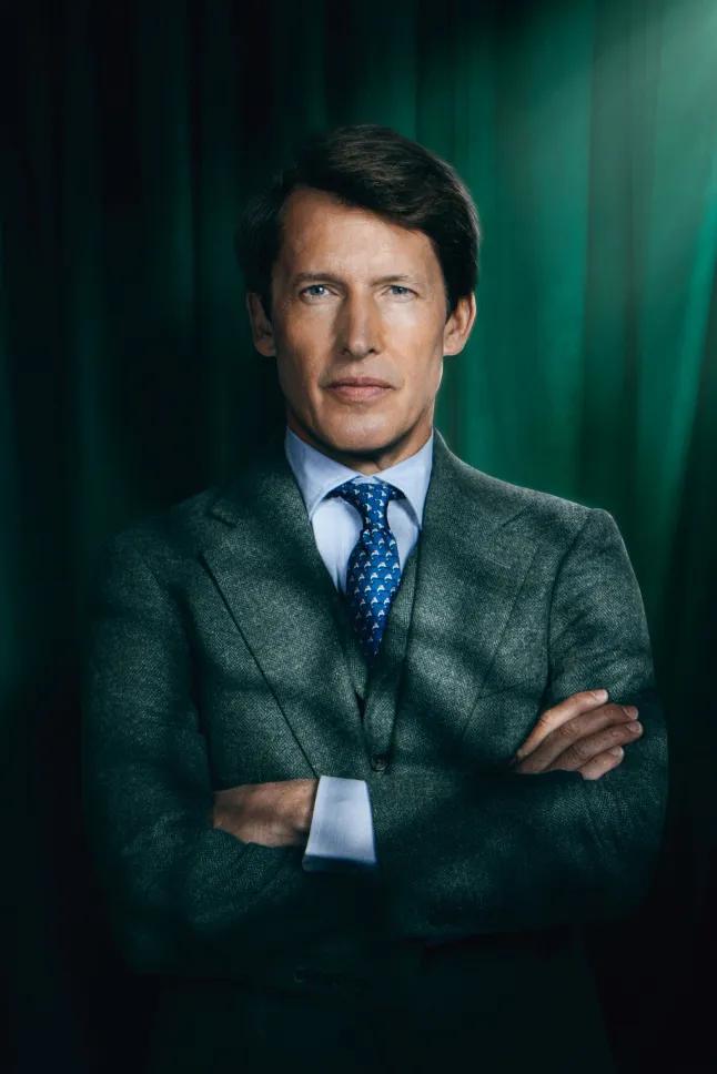 James Blunt in a green tweed suit, blue shirt and tie, arms folded in front of a green velvet curtain