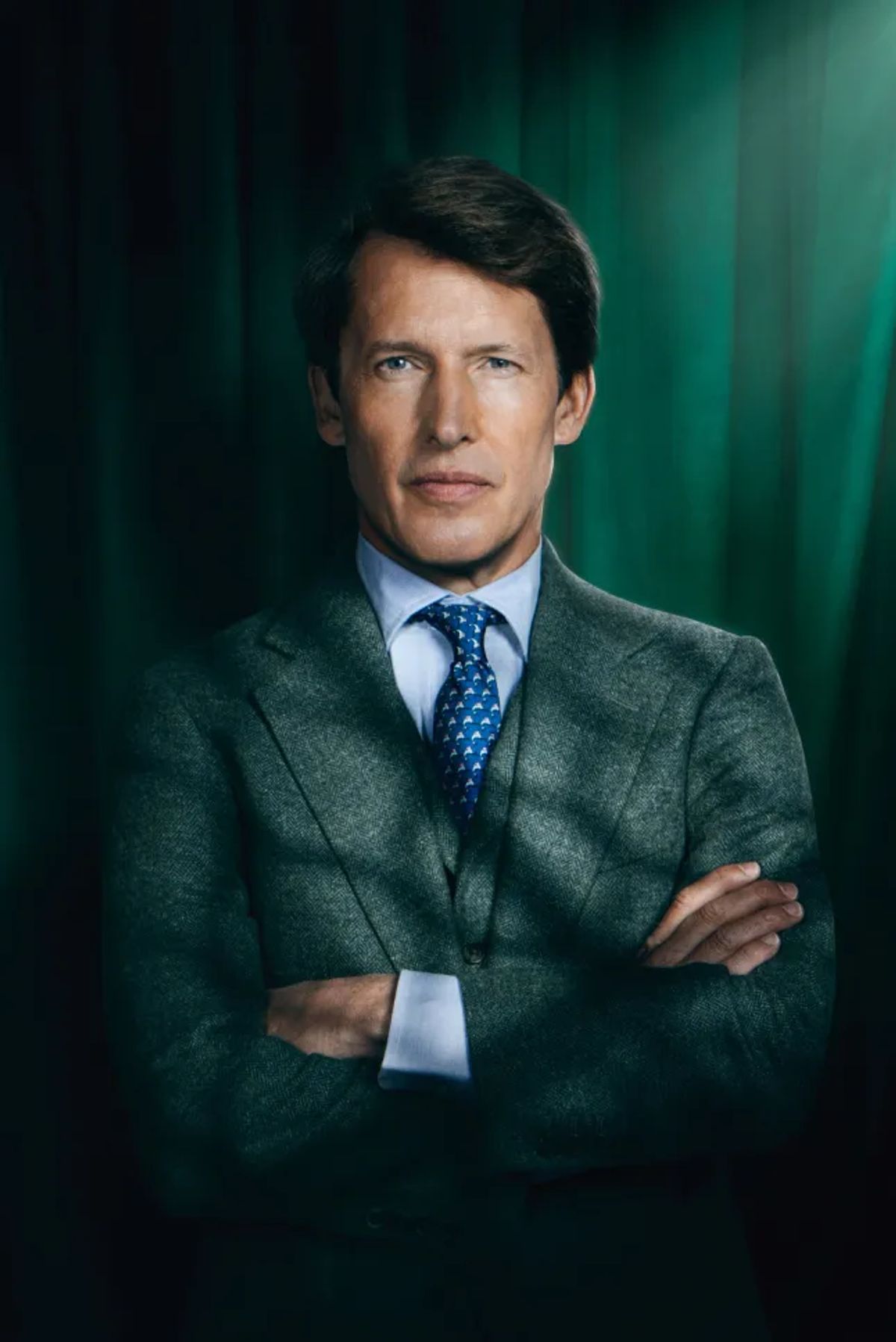 James Blunt in a green tweed suit, blue shirt and tie, arms folded in front of a green velvet curtain
