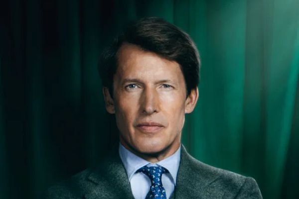 James Blunt in a green tweed suit, blue shirt and tie, arms folded in front of a green velvet curtain