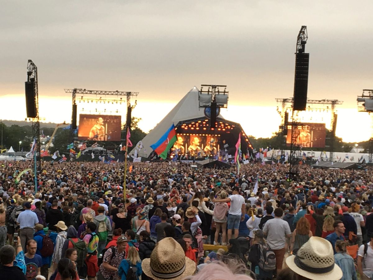 Huge crowd with flags facing the Pyramid Stage at dusk, with the act shown on big screens either side