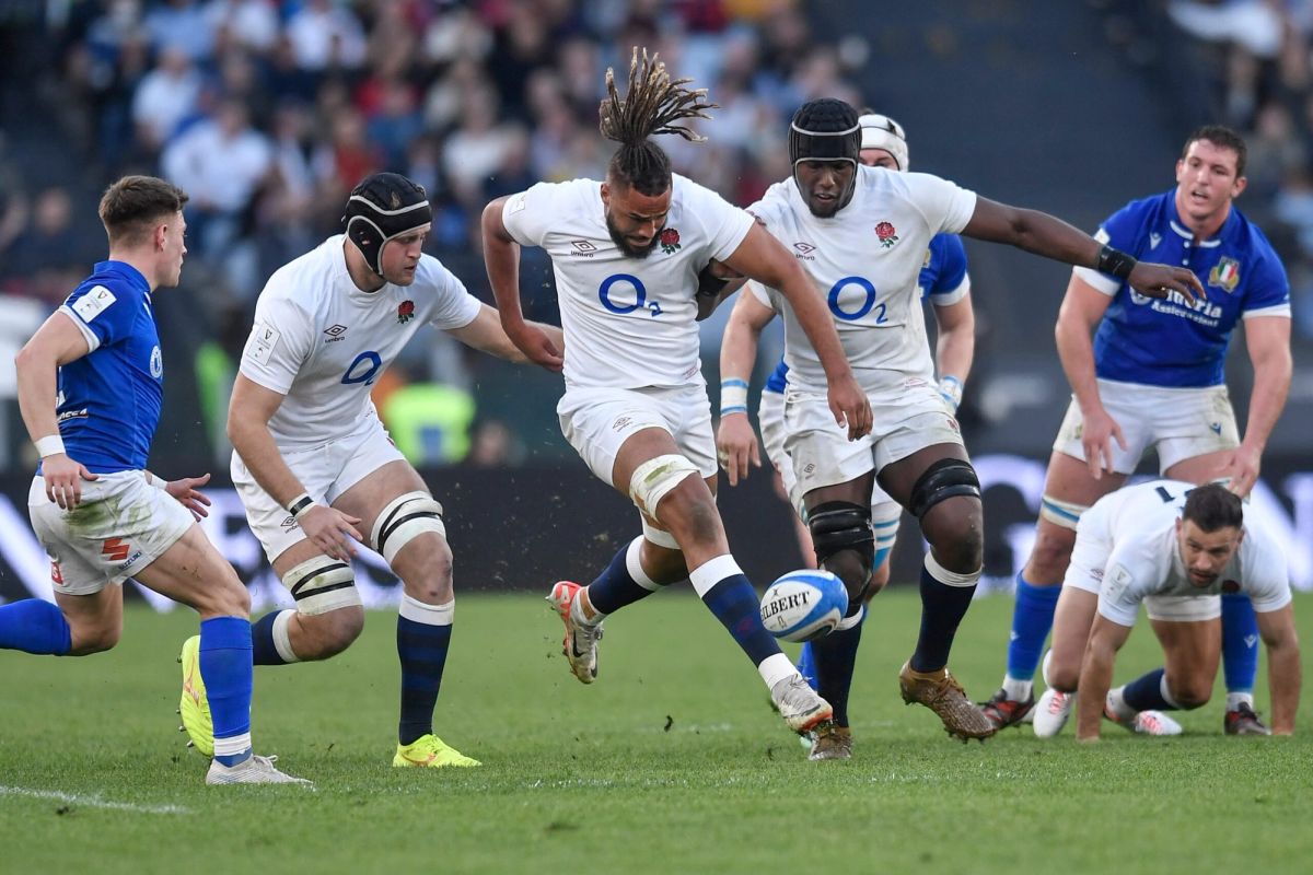 England's Fin Smith kicks rugby ball, dreadlocks flowing, as Italy players defend Six Nations.