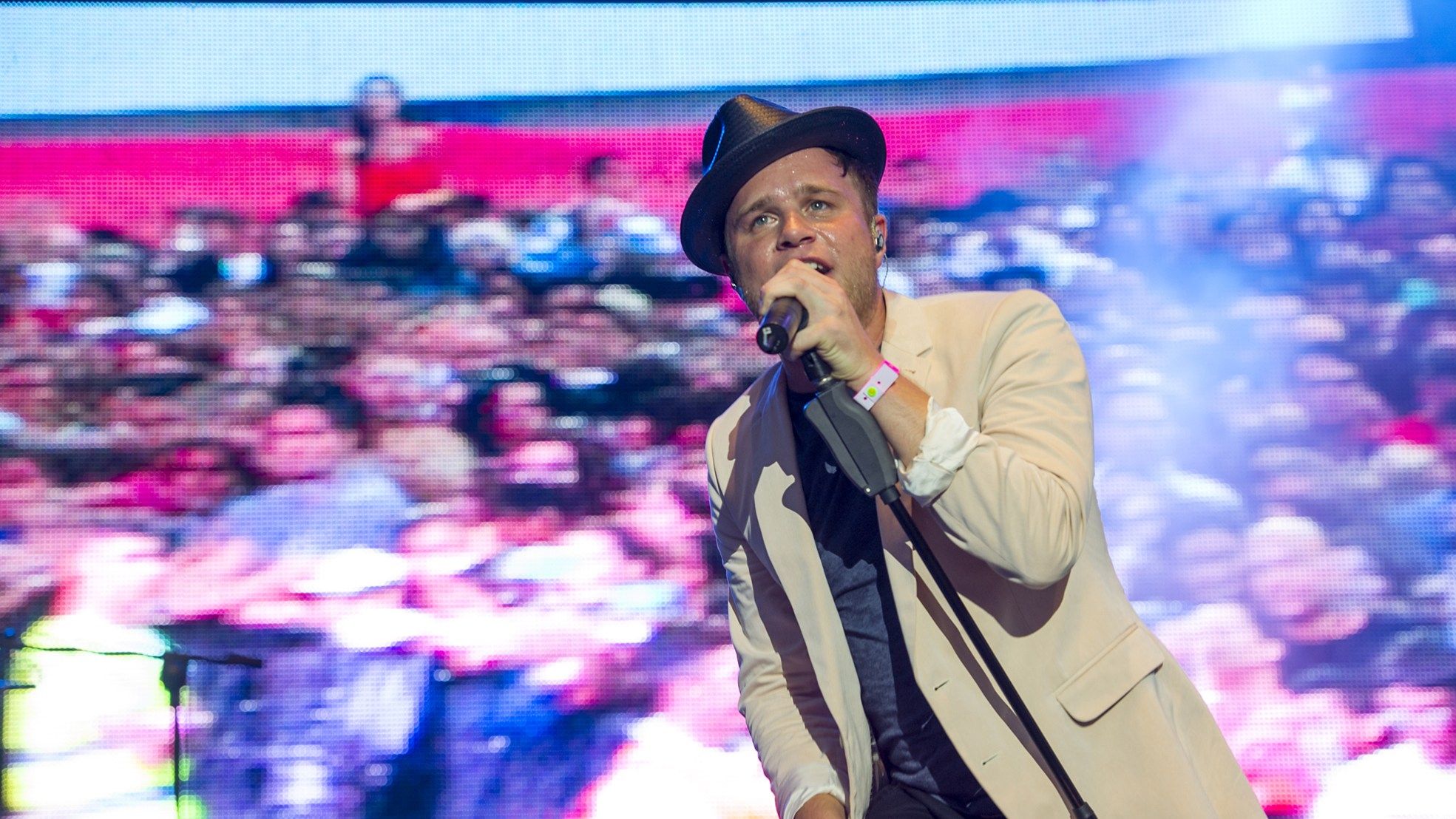 Olly Murs in a trilby and cream jacket singing in front of a big screen showing the crowd