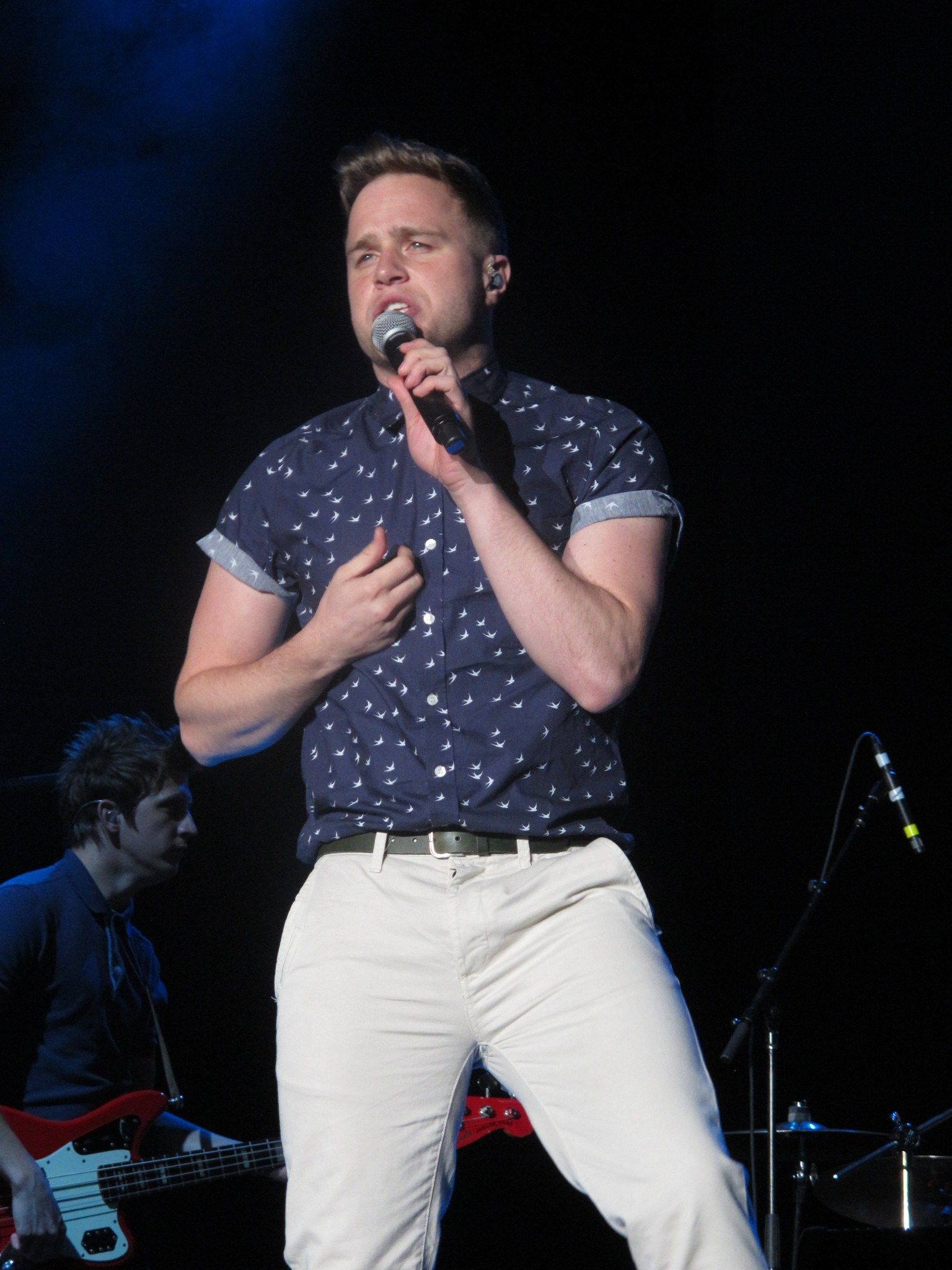 Olly Murs singing into a handheld microphone on a dark stage, with his bass player behind him