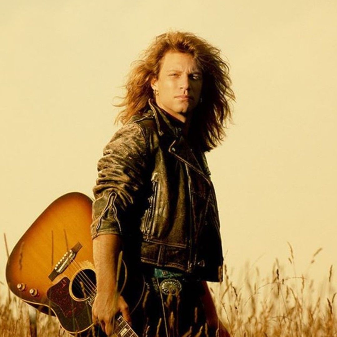 Jon Bon Jovi with long hair, leather jacket, holding an acoustic guitar in a golden field.