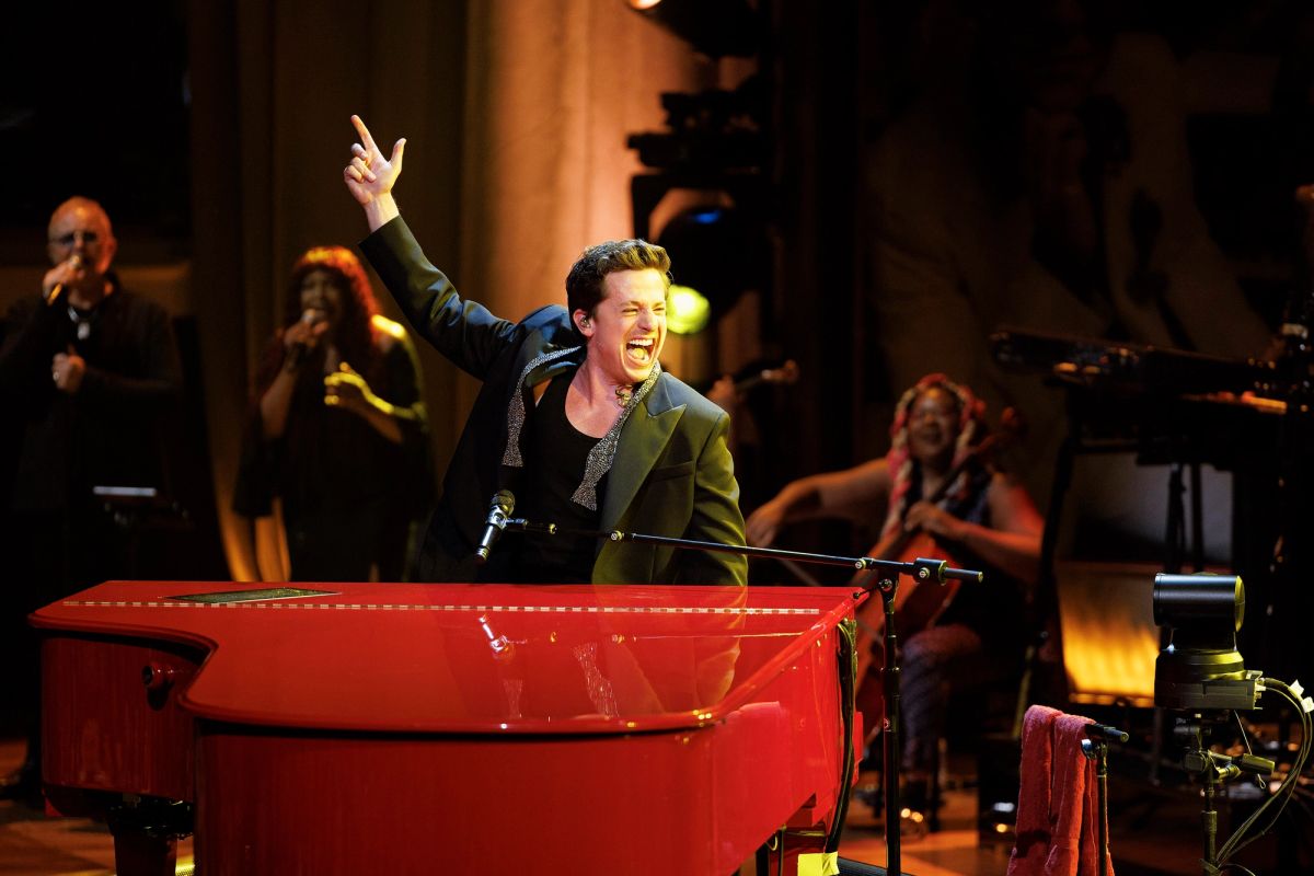 Charlie Puth at a red grand piano, pointing upwards and laughing, with backing singers behind him
