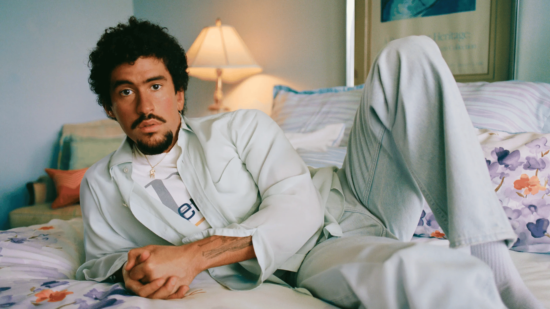 Artist reclines on bed in casual wear, blue wall with "Heritage" art. Relaxed bedroom portrait.