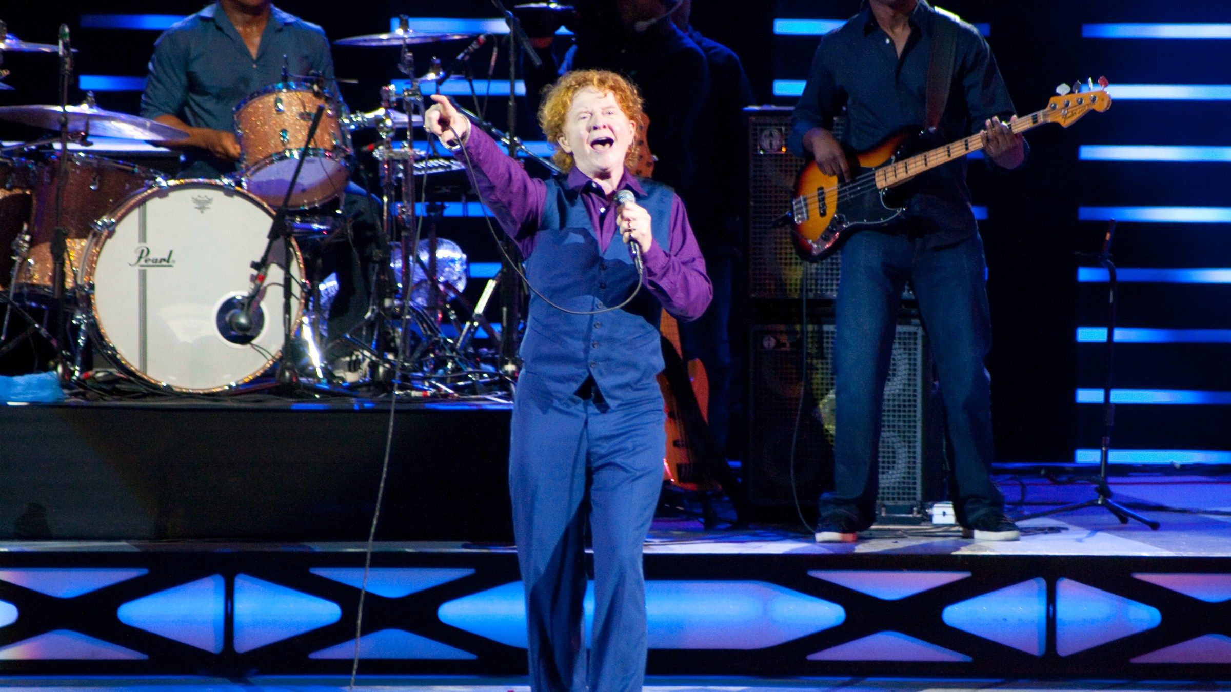 Mick Hucknall pointing to the audience as he sings, with the Simply Red drummer and bass player behind him
