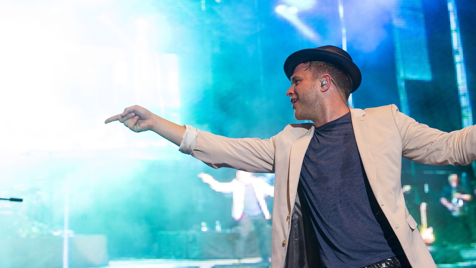 Olly Murs in a trilby and cream jacket pointing out to the crowd from a brightly lit stage
