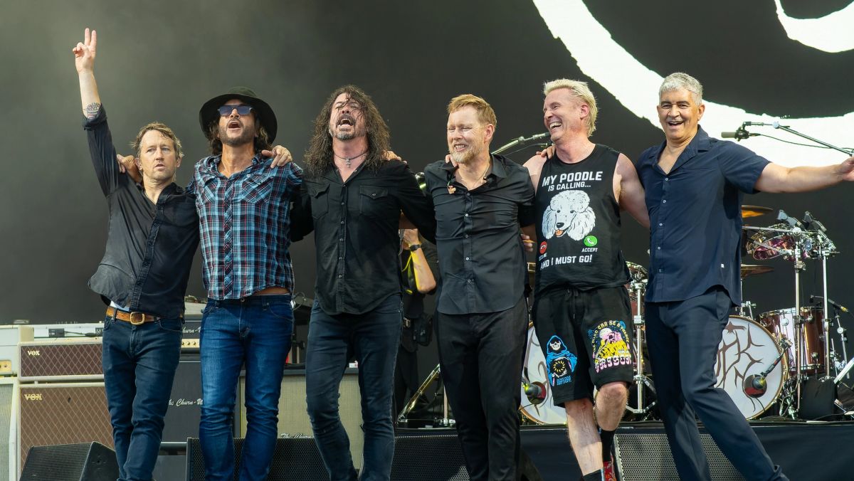 Six members of Foo Fighters stand arm in arm at the front of the stage, taking a bow in daylight