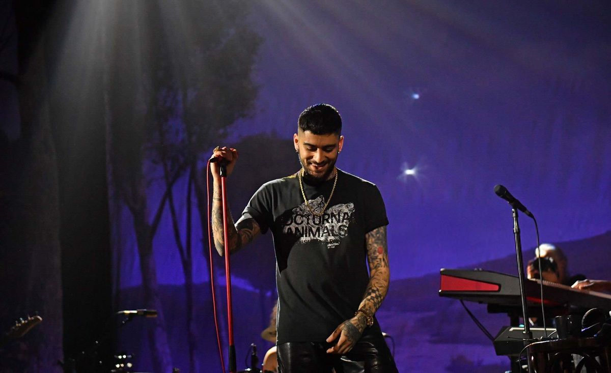 Zayn Malik sings on stage, "NOCTURNAL ANIMALS" shirt, tattoos, blue-purple lighting.