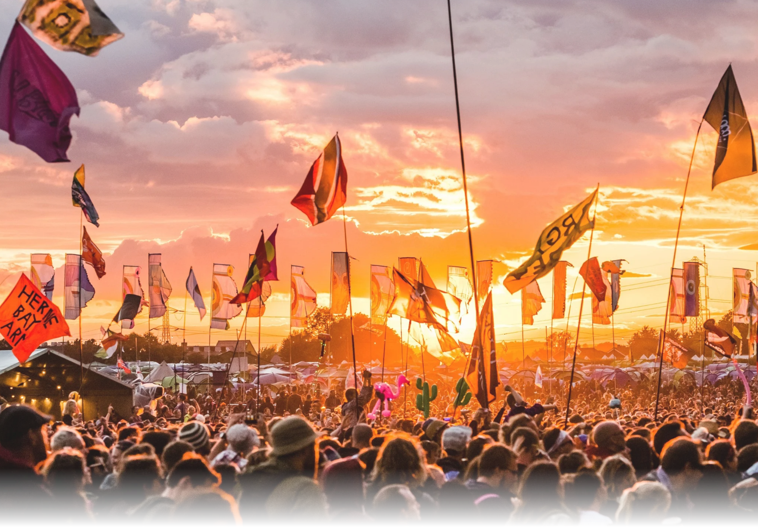Glastonbury crowd at sunset, flags aloft including BBC 6 Music, Herne Bay, and Boyce banners.